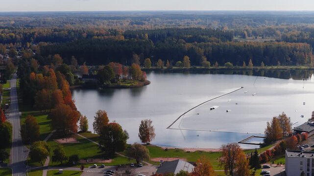 Aerial view of Jelgava, Latvia waterfront with promenade, sandy beach, pier, floating wakepark ramps and buoys, autumn foliage, soft daylight, and Jelgava Palace nearby.