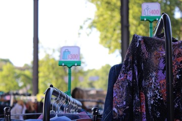 Feira de Moda Circular ao Ar Livre com Roupas em Araras, Clientes Desfocados ao Fundo e Placa de Promo&ccedil;&atilde;o Destacando Valor 300, Ambiente Natural