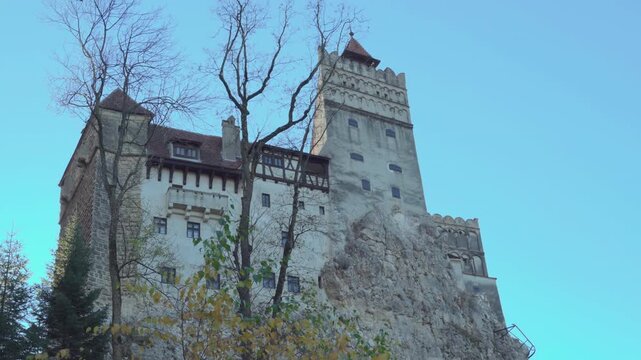 Historic Bran Castle, Romania low angle day exterior, perched on a rocky cliff surrounded by trees under a clear blue sky.