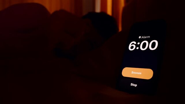 Man Lying in Bed Looking Toward Smartphone Alarm Ringing at 6 AM with Orange Light from Window Lazy Morning