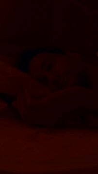 Vertical video. Man Lying in Bed Under White Sheets Turning Off 6 AM Alarm Looking Out the Window Then Rolling Over in Warm Dim Light Morning Laziness