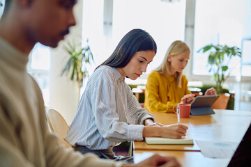 Young woman taking notes while collaborating with diverse colleagues in a modern office, focusing on teamwork and productivity