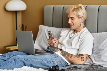 Young man enjoys leisure time on bed while checking phone and using laptop in modern apartment