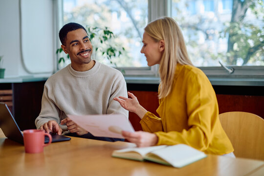 Two smiling business colleagues discussing documents and working on a laptop during an informal office meeting - Powered by Adobe