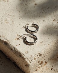 Pair of small silver hoop earrings rests upon a textured, light-colored stone surface with natural shadow patterns