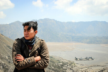 Pensive Asian Young Man on Mountain Ridge Overlooking Vast Volcanic Bromo. Conveying solitude, thought, and adventure.
