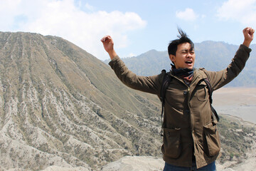 Excited Asian Young Man Hiker Celebrating Success on a Mountain Summit. Represents achievement, freedom, and the spirit of adventure.