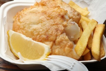 British Traditional Fish and chips with lemon in takeout container on wooden table, closeup