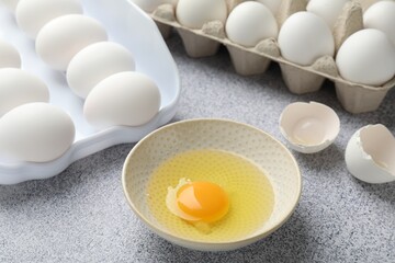 Many whole raw chicken eggs and broken one in bowl on grey table, closeup
