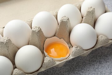 Many whole raw chicken eggs and broken one in carton on grey table, closeup