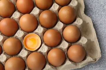Many whole raw chicken eggs and broken one in carton on grey table, top view
