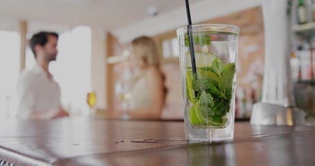 Showcasing mojito glass with mint, cucumber slices on bar counter, with blurred couple chatting