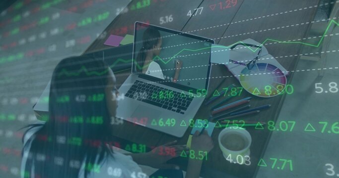 Analyzing stock charts African American woman taking notes at laptop in home office, with coffee