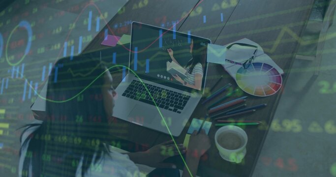 Watching student holding smartphone at home desk, with laptop, supplies, coffee mug, stock charts