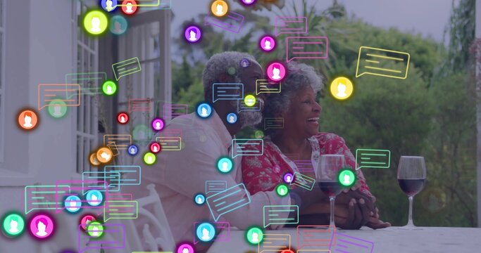 Smiling senior couple holding hands at patio table, with wine glasses and digital chat icons