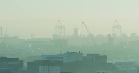 Towering container cranes rising above port city skyline, with warehouse rooftops and smog haze