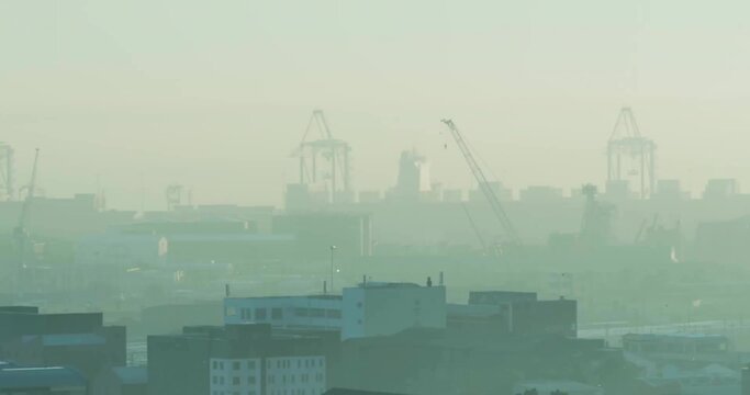 Towering container cranes rising above port city skyline, with warehouse rooftops and smog haze
