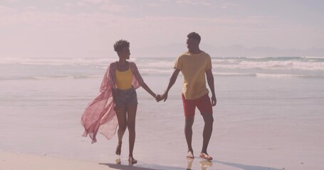 Walking barefoot couple wearing pink shawl, tank top, swim shorts on wet sand with gentle waves