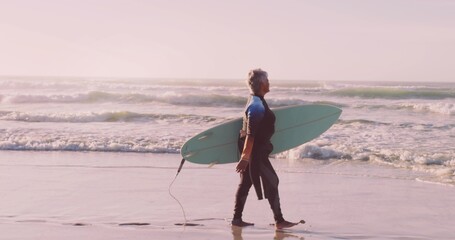 Walking senior woman carrying light green surfboard along sandy shoreline, with neoprene wetsuit