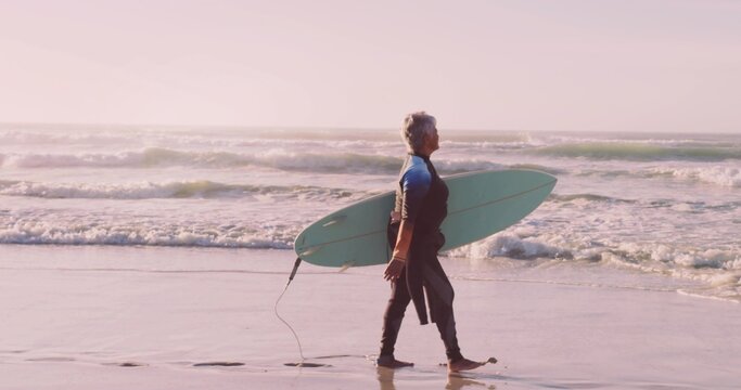 Walking senior woman carrying light green surfboard along sandy shoreline, with neoprene wetsuit - Powered by Adobe