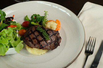 A top-down shot of a gourmet steak on a plate alongside fresh vegetables, with cutlery. The steak is grilled perfectly and served on a bed of mashed potatoes