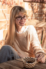 Smiling woman with glasses sitting at a rustic table with a coffee cup in natural light.