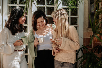 Three women laughing together while sharing coffee in a cozy indoor garden setting filled with...