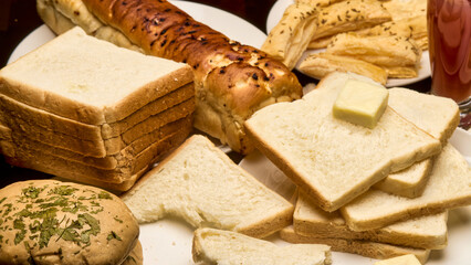 A beautifully arranged assortment of bakery items featuring soft white bread slices topped with butter, a loaf of garlic bread, crispy puff sticks, and a glass of fruit juice captured in warm light