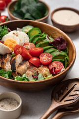 Healthy Cobb salad with grilled chicken breast, avocado, cherry tomatoes, lettuce and boiled egg in wooden bowl, top view on light background