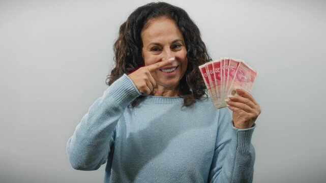 Middle-aged woman holding israeli shekel banknotes gives a thumbs-up sign against a white background, emphasizing wealth and contentment.