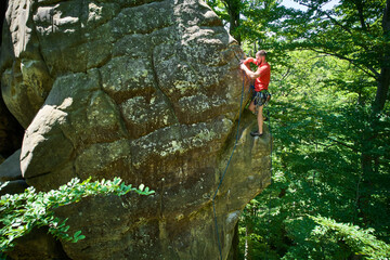 Aerial view of male rock climber ascending rugged limestone cliff with harness and rope for safety. Sportsman climbing on vertical large boulder at Dobvush Rocks in Carpathian mountains, Ukraine.