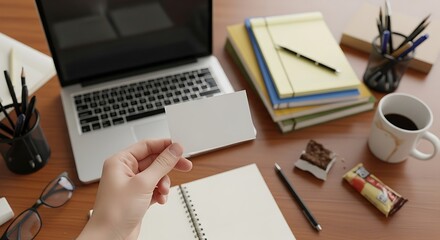 Top view of a person holding a blank business card next to a laptop, notebook, and coffee cup on a wooden desk, creating a workspace scene