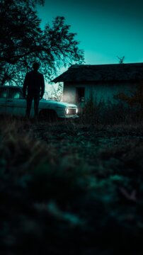 Silhouetted figure in eerie twilight by vintage car, embodying cryptic suspense and Samhain's haunting whispers with shadowed intrigue