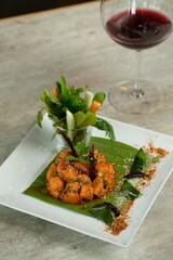 A gourmet meal featuring cooked shrimps on a banana leaf and a glass of red wine. Fresh vegetables are placed separately. The scene is set with style and class.