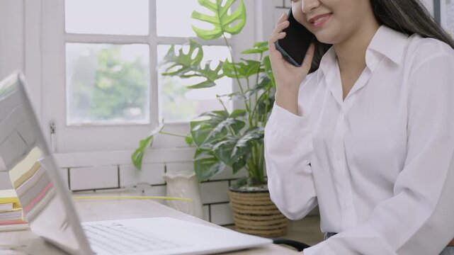 Closeup of happy young Asian female teacher sitting smiling and typing keyboard on laptop computer and using her mobile phone technology talking at her desk in classroom. Back to school concept. 