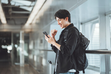 Happy young Asian traveler celebrating success while looking at smartphone in airport terminal,...