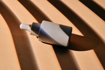 A bottle of serum on a beige background with stripes of shadows.