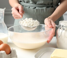Making batter (liquid dough). Woman sieving flour into bowl at white table indoors, closeup
