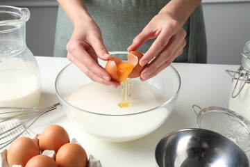 Making batter (liquid dough). Woman adding eggs into bowl at white table indoors, closeup