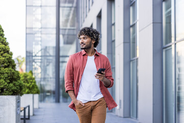 A young Indian man in a red shirt is walking down the street, holding his hand in his pocket, using his phone, and looking to the side with a smile