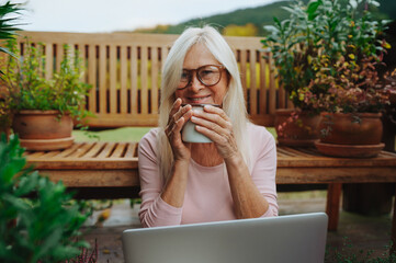 Older woman having cup of tee while working from homeoffice, sitting on terrace.