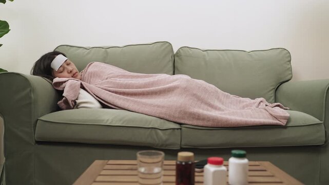 Sick young Asian woman feeling unwell and having a headache using a fever cooling patch on her forehead to relieve symptoms and lying covered up with a blanket on sofa in the living room at her home. 