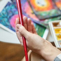 Female artists hand holding red brush with colorful paintings and watercolor set in background.