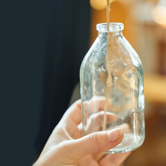 Female hand holding glass bottle with plant stem indoors in soft focus lighting.