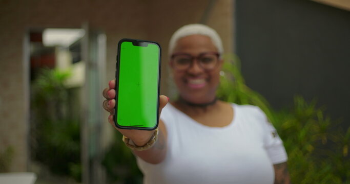 African American woman holding smartphone forward, green screen Chroma Key on display, focus on device with blurred smiling face in background, modern technology and digital connection concept - Powered by Adobe