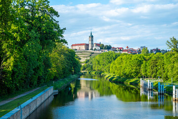 Naklejka premium Landscape of Melnik with a calm rive with historic castle with a church tower. The scene is bright and peaceful, with clear skies and soft clouds adding to the charm of the view.