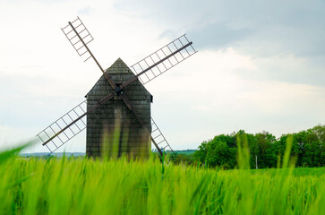 Old wooden windmill standing in a green field of tall grass with trees and cloudy sky in the background. Traditional rural landscape.