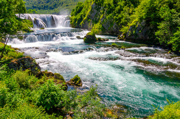 Beautiful turquoise river and waterfall surrounded by lush green forest in sunny summer landscape