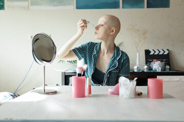 Bald woman after chemotherapy applies skincare serum in front of a mirror at home, representing...