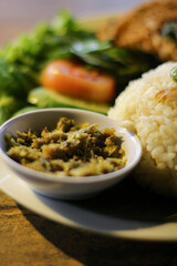 Traditional Indonesian Nasi Liwet served on a wooden table. The dish includes steamed rice, fried tempeh or egg topping, vegetables, sambal, and herbal tea.